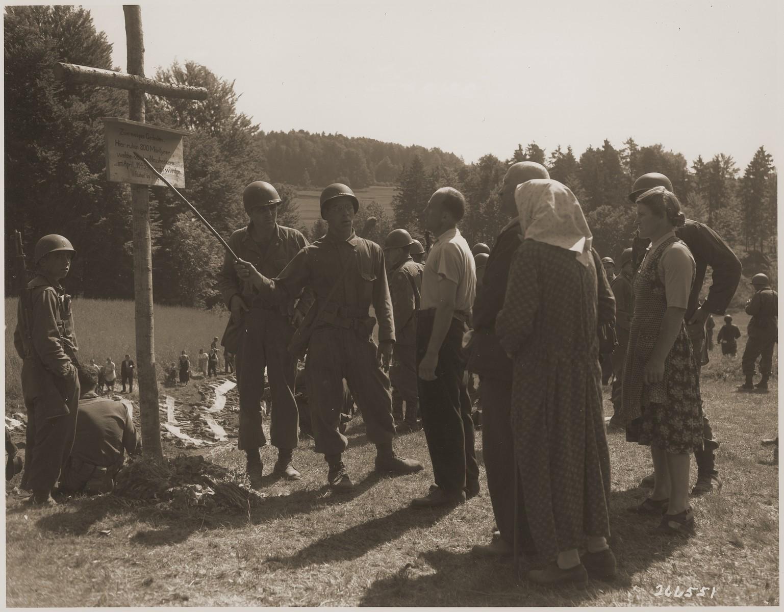 An American soldier pointing out a sign to German civilians that memorializes the 800 prisoners shot by the SS near the town of Nammering, 1945. In the background, other German civilians walk among corpses exhumed from the mass grave. (1537×1200)