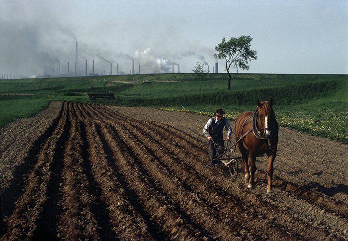 Polish man using a horse-drawn plow to tend to his field, as smoke rises from industrial factories in the distance, Poland, c. 1981. – photo taken by French-Swiss photographer Bruno Barbey. [700 x 485]