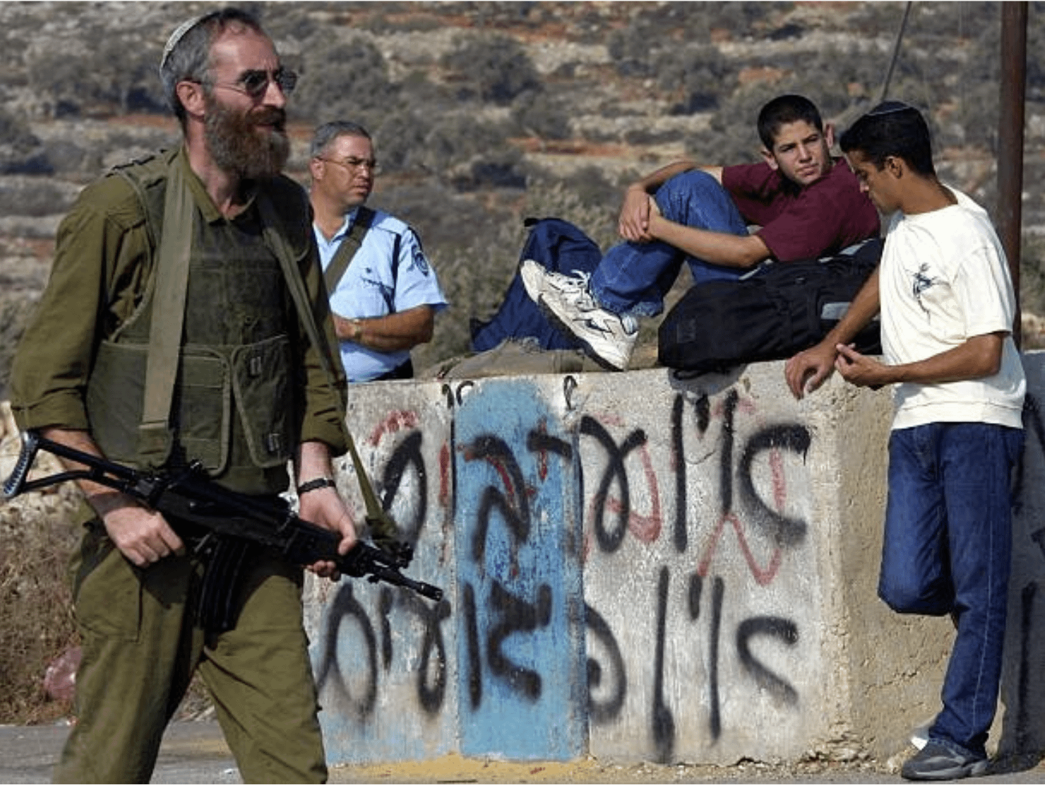 Young Israeli settlers rest on concrete blocks sprayed with grafitti that reads “No Arabs No Terror” as they wait for a bus, guarded by soldiers at Tapuah Junction checkpoint on the southern outskirts of the West Bank city of Nablus, 25 September 2001. [1480 x 1112]