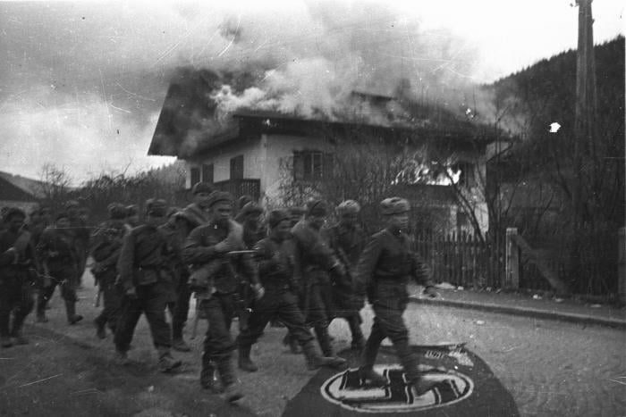 Soviet troops trample a Nazi flag as they march past a burning house on a street in the outskirts of Vienna, Austria, April 1945 [700×467]