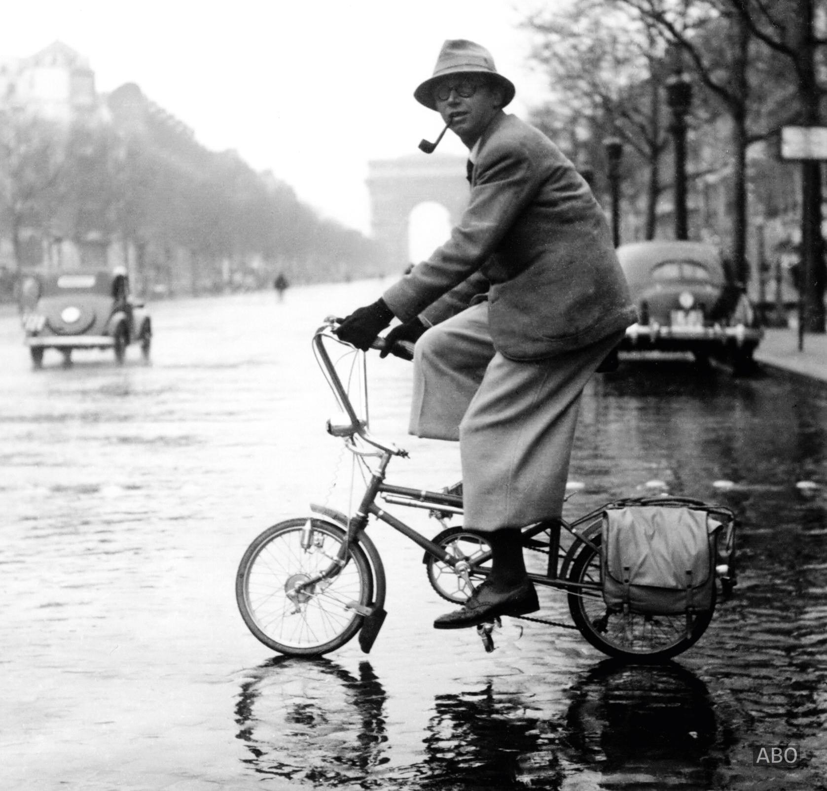 85 years ago… “Philosopher Jean-Paul Sartre (1905 – 1980) cycling on the rainy Champs-Élysées”; France – Paris, 1941 (WW2) [1677×1605]