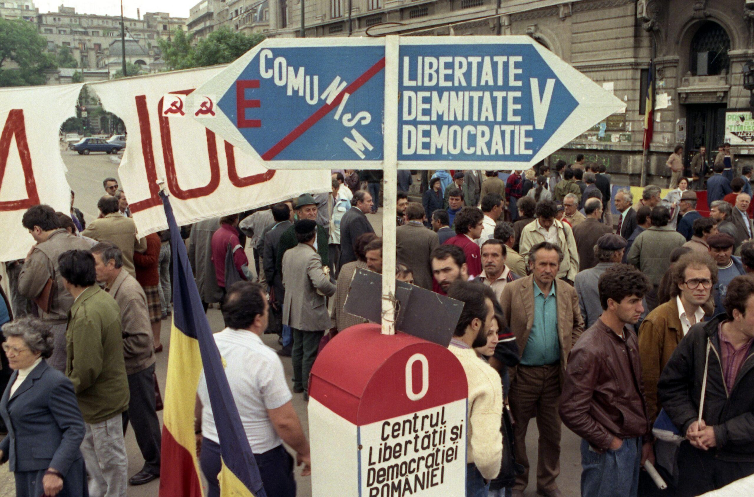 Demonstrators gathered at University Square in Bucharest, Romania, near a protest sign symbolizing the choice between Communism and the values of Liberty, Dignity, and Democracy, months after the fall of the Ceaușescu regime. (1990) [3000×1976]