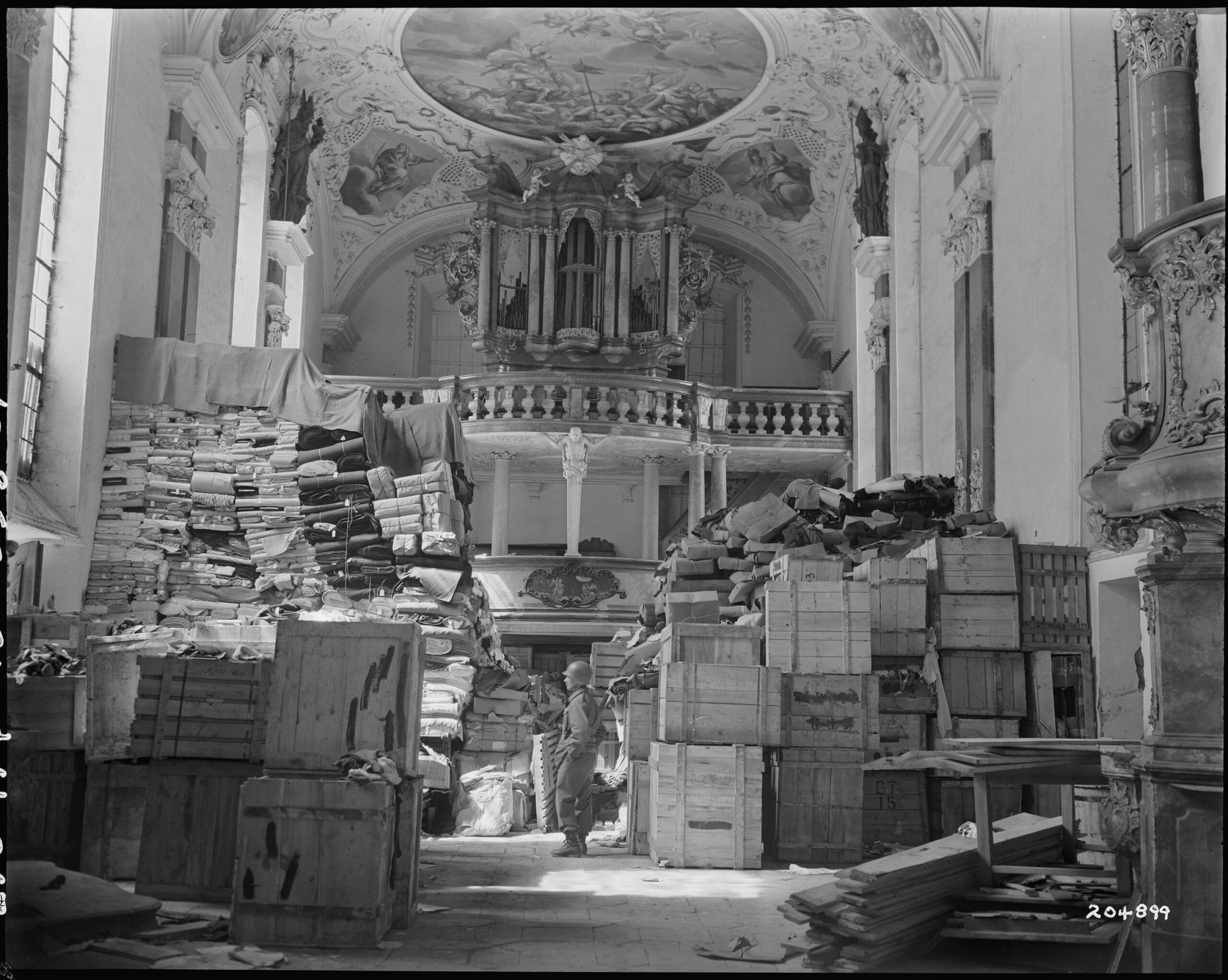 A US soldier inspecting piles of looted art and goods being stored in a Bavarian church at the end of WWII. (1945) [3006×2400]