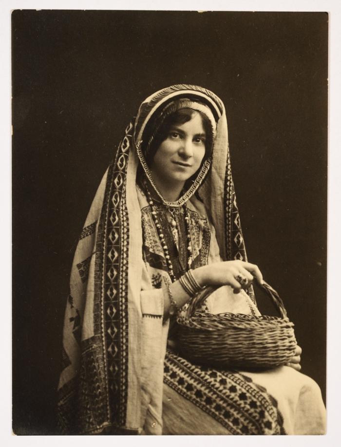 A Palestinian woman sitting with a straw basket in her hand wearing a Palestinian dress and a traditional shutwa headdress for the Ramallah area, 1930 [760×1000]