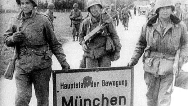 GI’s holding the sign marking the entrance to Munich with a bullet hole, which reads “Capital of the Movement“, 1945, 606 × 341