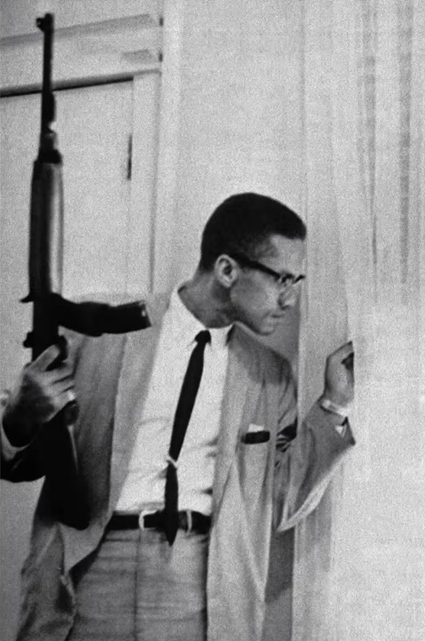 Malcolm X holding an M1 Carbine and pulling back the curtains to peer out of a window. (1965) [1439×2170]